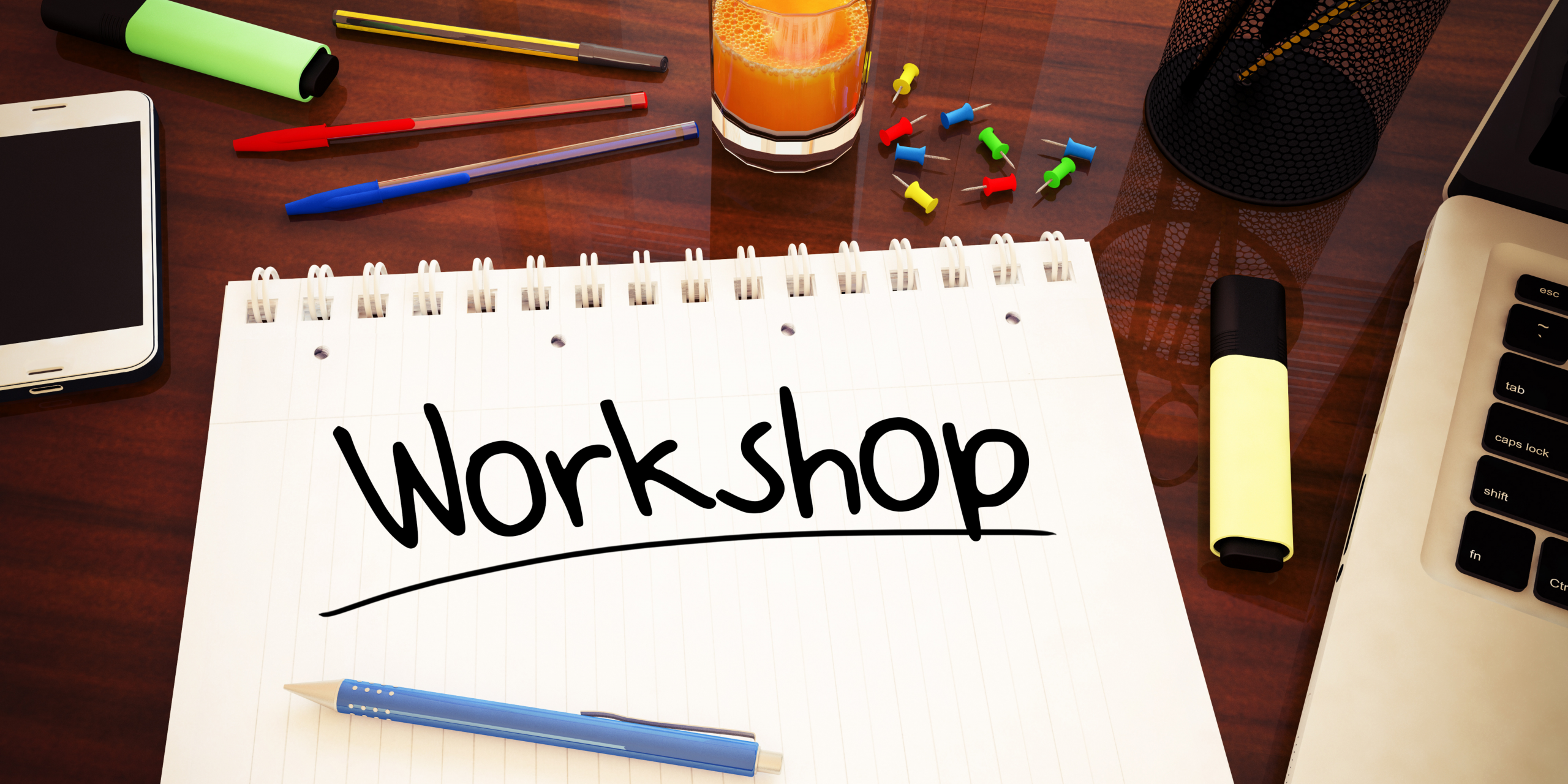 Photo of a desk surface with an opened notepad with "Workshop" written on and some pens, pencils, coloured highlighters, a mobile phone & the edge of a laptop.